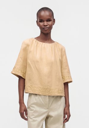 Femme aux cheveux courts portant un haut beige ample avec des bords brodés et un pantalon beige clair, debout devant un fond uni.