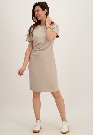 Vrouw die een beige jurk tot op de knie draagt met korte mouwen, witte sneakers en gouden oorringen, staand tegen een effen lichte achtergrond.