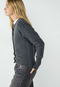 Dunkelgrauer gestrickter Cardigan mit langen Ärmeln, geripptem Saum und Knöpfen vorne. Kombiniert mit dunklen Jeans, die seitliche Taschen haben.