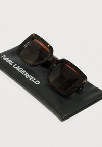 Színtarka napszemüveg négyszögletes kerettel fekete tokon. A tokon fehérrel a "KARL LAGERFELD" felirat olvasható. A lencsék sötétek és tónusosak.