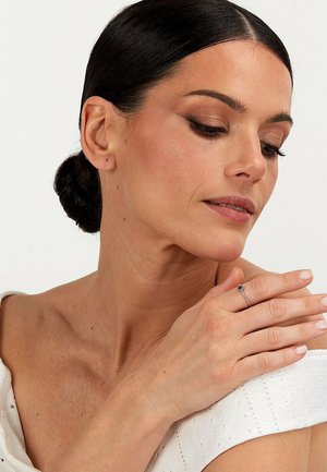 Silberner Ring mit einem kleinen blauen Stein, getragen an den Fingern, vor einem strukturierten weißen Stoffhintergrund. Das Model hat ein dezentes Make-up und dunkles Haar.
