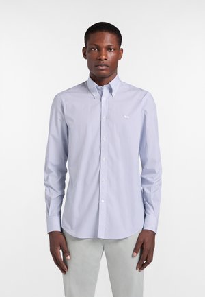 Junger Mann, der ein hellblaues, gemustertes Hemd mit Knöpfen und hellgraue Hose trägt, vor einem schlichten weißen Hintergrund stehend.