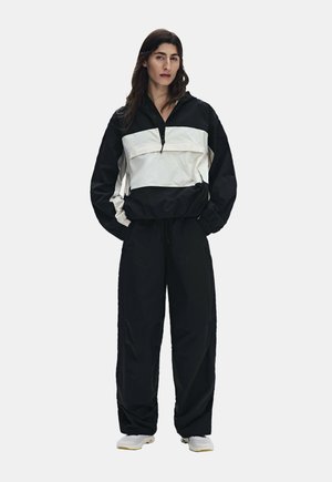 Kvinde med langt mørkt hår iført en sort-hvid pulloverjakke, sorte løse bukser og hvide sneakers, står med hænderne i lommerne.