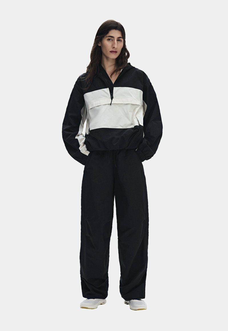Femme aux longs cheveux foncés portant une veste-pull noire et blanche, un pantalon noir ample et des baskets blanches, debout les mains dans les poches.