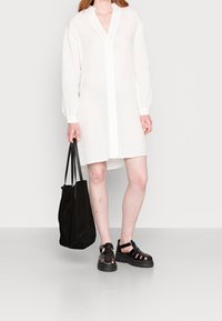 Bílá lněná košilová šaty s dlouhými rukávy a výstřihem do V, doplněné černými sandály na pásky a černou tote taškou. Hladká textura, minimalistický design.