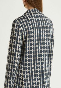 Blazer à motif géométrique bleu marine et beige, coupe ajustée, col à revers crantés, et texture lisse. Comprend des poches latérales.