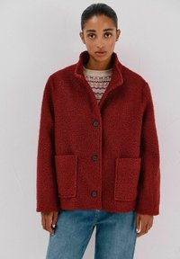 Rote, flauschige Jacke mit einem Kragen, zwei Fronttaschen und schwarzen Knöpfen. Getragen über einem gemusterten Pullover und kombiniert mit blauen Jeans.