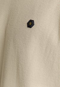 Beige textiel met een gebreide structuur en een hexagonaal zwart patch met gele accenten, dat een eenvoudig en casual ontwerp toont.