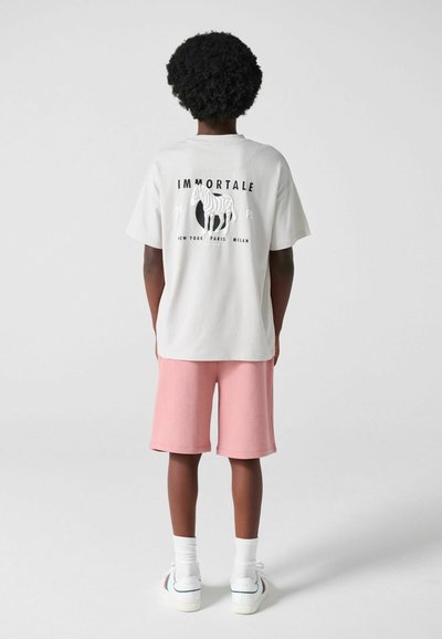 Barn med krøllet hår iført en hvid T-shirt med et zebra-grafik og teksten "IMMORTALE", lyserøde shorts, hvide sokker og hvide sneakers, står med ryggen til.
