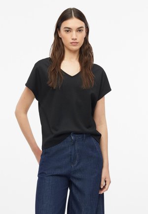 Giovane donna con lunghi capelli castani che indossa una maglietta nera a maniche corte con scollo a V e jeans blu a vita alta e gamba larga, in piedi davanti a uno sfondo bianco.