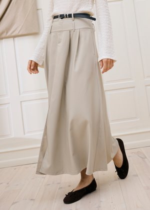 Femme portant un pantalon beige à jambes larges avec une ceinture noire, un pull blanc texturé et des ballerines noires, marchant à l'intérieur sur un sol en bois clair.