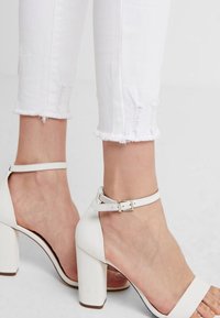 Sandalias de tacón bloque blanco con material texturizado, correa en el tobillo y detalle de hebilla. Combinadas con jeans blancos desgastados y con dobladillo.