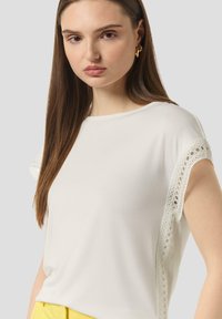 White short-sleeve top featuring lace detailing along the sleeves. Soft fabric with a relaxed fit, worn with yellow bottoms for contrast.