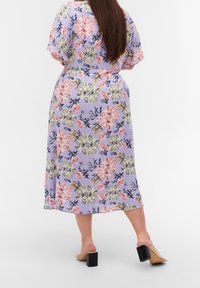 Blommig klänning med korta ärmar, lavendelgrund, stort mönster av rosa och gula blommor, medellång, kompletterad av beige öppna tofflor.