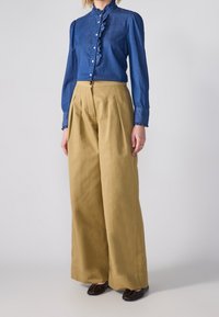 Jeansskjorta med volangkrage och långa ärmar, tillsammans med högmidjade, veckade khakibyxor och mörkbruna lägenhetsskor.