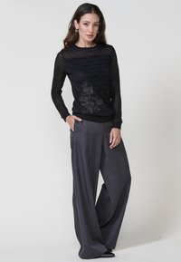 Una donna sta in piedi con sicurezza, indossando un maglione nero con dettagli in pizzo e pantaloni grigi larghi, in posa contro uno sfondo semplice.