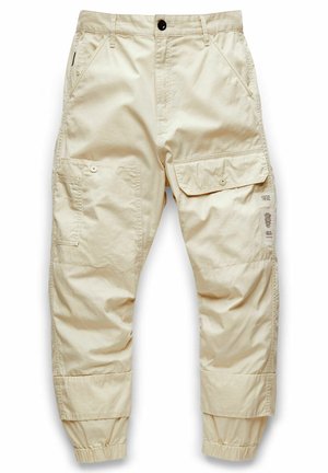 Pantalon cargo beige avec rabat boutonné et poches latérales, poignets élastiques, passants de ceinture et fermeture avant par boutons, présenté sur fond blanc.