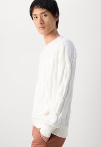 Nuori mies, joka on pukeutunut valkoiseen nysväneuleeseen ja ruosteenvärisiin housuihin, seisoo yksi käsi taskussa yksinkertaista valkoista taustaa vasten.