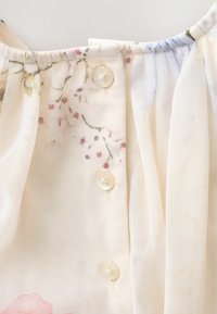 Leichtes, durchsichtiges Oberteil aus Stoff mit Blumenmuster. Verfügt über einen gerafften Ausschnitt und drei cremefarbene Knöpfe auf der Vorderseite. Weiche Textur.