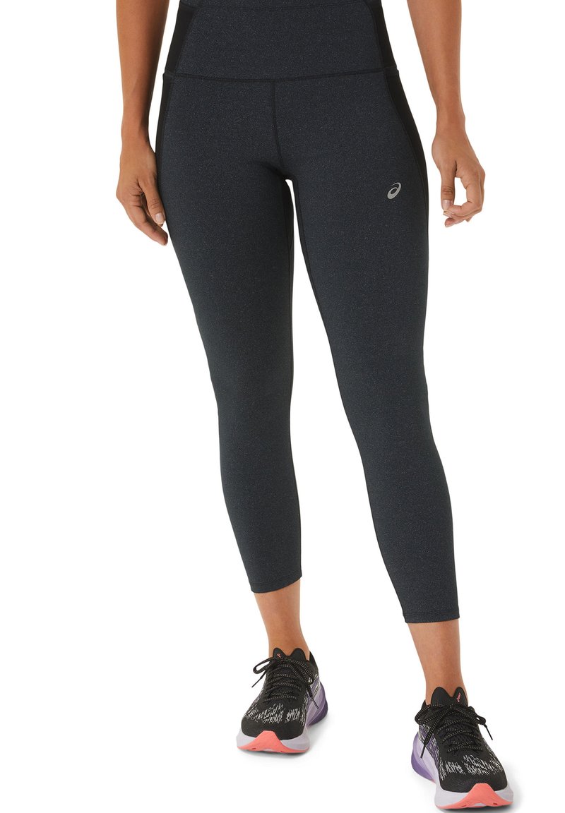 Mujer vestida con leggings deportivos negros y zapatillas de correr negras con detalles en rosa y púrpura, de pie con los brazos relajados a los lados.