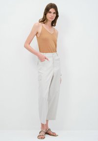 Tans top, lichtgrijze brede broek met zijzakken en sandalen met gouden accenten, model staat tegen een plain witte achtergrond.