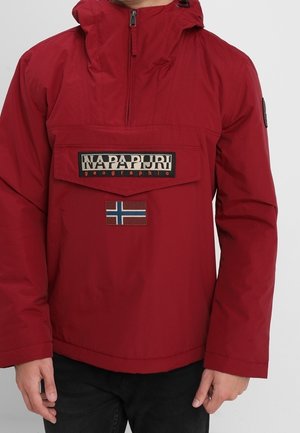 Veste rouge à capuche avec rabat de poche avant affichant le logo "Napapijri geographic" et un écusson du drapeau norvégien en dessous, portée avec un pantalon noir.