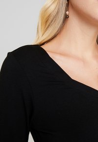 Musta V-pääntie paita, pehmeästä ja joustavasta materiaalista. Lähikuva olka-alueesta, joka näyttää sileän pinnan ja siistit saumat. Yksinkertainen muotoilu.