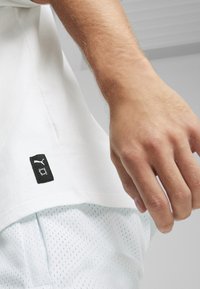 Biela bavlnená tričko s čiernou logom nášivkou na leme. Viditeľná ruka má svetlú pleť a je umiestnená na svetlých sieťovaných šortkách.