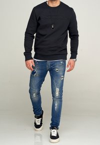 Felpa nera con logo "JACK&JONES" in rilievo, abbinata a jeans blu strappati con buchi e sneakers nere con dettagli bianchi.