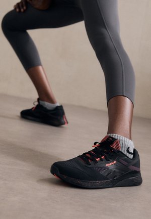 Persona con leggings grigi che indossa sneakers nere e arancioni Reebok, in posizione di affondo con gambe larghe su un pavimento liscio.