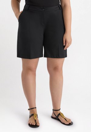 Pantaloni corti neri sartoriali con una texture liscia, caratterizzati da tasche laterali e chiusura frontale con bottone. Indossati con sandali neri con accenti dorati.