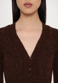 Gros plan sur une personne portant un pull en maille texturée brun foncé avec un col en V profond et une petite fermeture éclair dorée au centre devant.