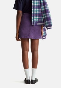 Mini falda de pana morada con bolsillos traseros, combinada con calcetines blancos y zapatos negros. Una chaqueta de cuadros cuelga de un hombro.