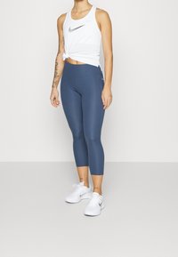 Biały top na ramiączkach z wiązanym detalem i srebrnym logo Nike, zestawiony z granatowymi legginsami 3/4 oraz białymi butami sportowymi.