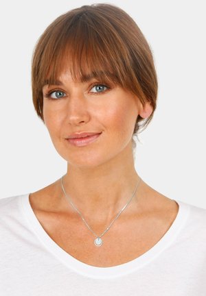 Collana d'argento con pendente circolare, indossata su un collo nudo, con una camicia bianca semplice. Il tono della pelle appare chiaro, i capelli sono castani e di lunghezza spalle.