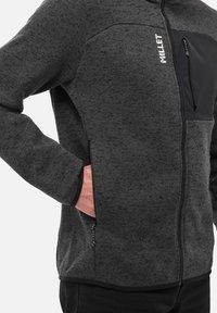 Veste en polaire texturée noire et grise avec une poche zippée sur la poitrine, des poches avant et des bordures noires contrastées le long des poignets.