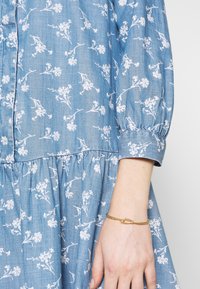 Jasnoniebieska sukienka z chambray z białym kwiatowym wzorem, długimi rękawami bufkami i marszczoną talią; noszona z złotą bransoletką.