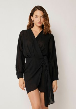 Femme aux cheveux bruns ondulés, portant des boucles d’oreilles créoles dorées et une robe portefeuille noire à manches longues sur un fond uni.