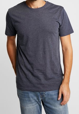 Homme portant un t-shirt à manches courtes gris foncé uni et un jean bleu clair, debout avec une main détendue le long du corps.