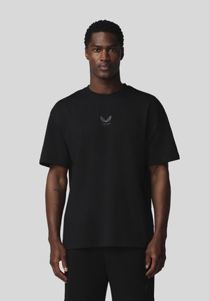 Homme aux cheveux courts et bouclés portant un t-shirt noir avec un petit logo blanc ailé et un pantalon noir devant un fond gris uni.
