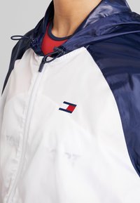 Giacca a vento bianca e navy con chiusura a zip, cappuccio regolabile e un piccolo logo con accenti rossi sul petto. Materiale liscio e leggero.