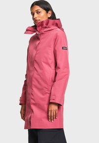 Femme portant un long imperméable rose avec un col montant et une capuche, debout devant un fond clair uni.