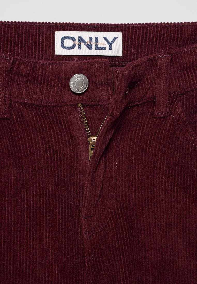 Bordeaux corduroy broek met een ritssluiting, metalen drukknoopsluiting en ribbeltextuur. Het label zegt "ONLY" in het blauw.
