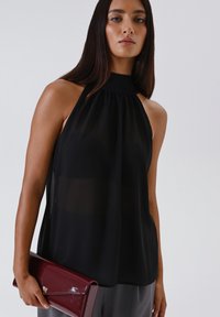 Top a halter nero realizzato in tessuto trasparente con collo alto. La modella tiene una pochette bordeaux lucida, che completta i pantaloni grigi.