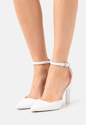 Des pieds portant des escarpins blancs à bout pointu avec des brides de cheville et des boucles, sur un fond clair et uni.