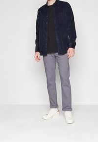 Námořnická manšestrová košile, černé tričko s kulatým výstřihem, šedé kárované kalhoty a bílé tenisky s barevnými akcenty. Model stojí s uvolněnýma rukama.