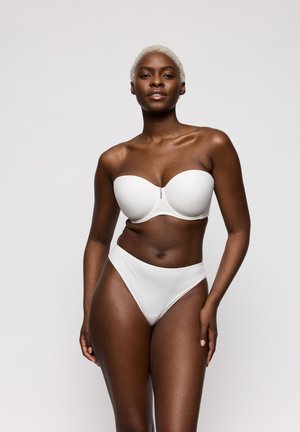 Zwarte vrouw met kort blond haar, gekleed in een witte strapless bh en bijpassend hoog uitgesneden ondergoed, staand tegen een effen lichte achtergrond.