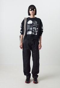 Felpa nera con stampe grafiche bianche, abbinata a pantaloni cargo neri larghi. Il modello indossa occhiali da sole e sneakers sportive nere/rosse.