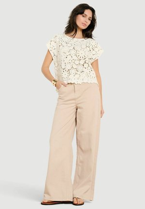 Žena oblečená v bielom háčkovanom tričku s krátkym rukávom, béžových nohaviciach s širokými nohavicami, sandáloch a náramku, pózujúca s jednou rukou v vrecku.