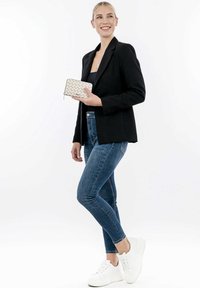 Black blazer over a fitted top, paired with blue jeans and white sneakers. Holding a geometric-patterned cream wallet. Simple background.